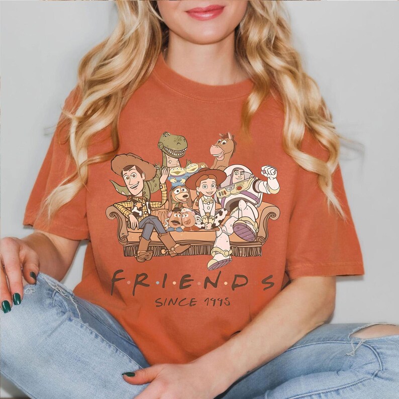 May include: Burnt orange t-shirt featuring a graphic of Toy Story characters on a couch. The word "FRIENDS" is printed below the graphic, with "SINCE 1995" underneath. The shirt is made of a soft material.