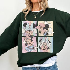 May include: Dark green sweatshirt featuring a four-panel design with Minnie Mouse illustrations. Each panel showcases a different pose of Minnie Mouse, with pastel-colored backgrounds and a white collar.
