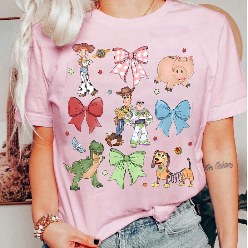 May include: Pink t-shirt featuring cartoon characters and bows. The design includes images of a pig, a dinosaur, a dog with a spring body, and other characters. The shirt also has several bows in different colors.