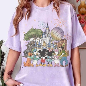 May include: A light purple t-shirt featuring a colorful graphic of a theme park with a castle, train, and characters like Mickey and Minnie Mouse. Fireworks are in the background.