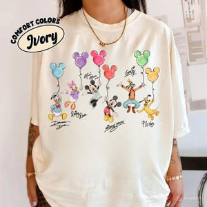 May include: Ivory t-shirt with Disney characters and Mickey Mouse-shaped balloons. Characters include Donald Duck, Daisy Duck, Mickey Mouse, Minnie Mouse, Goofy, and Pluto. The shirt has the words "Comfort Colors" and "Ivory" printed on it.