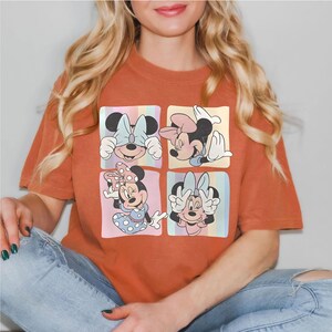 May include: Burnt orange t-shirt featuring a four-panel design of Minnie Mouse in various poses. Each panel has a pastel striped background. Minnie is wearing different colored bows and outfits. The shirt is made of a soft material.