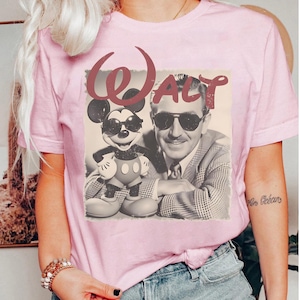Puede incluir: Camiseta rosa claro con un gr&aacute;fico retro de Mickey Mouse con gafas de sol y un retrato de Walt Disney. La palabra "Walt" est&aacute; escrita encima de la imagen en una fuente estilizada. La camiseta tiene mangas cortas.