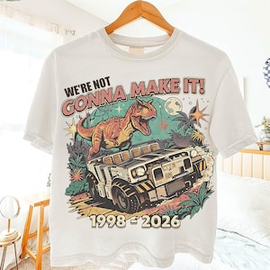 Op de afbeelding: Wit T-shirt met een kleurrijke afbeelding van een dinosaurus, een jeep en de tekst "WE'RE NOT GONNA MAKE IT!" en "1998-2026". Het ontwerp bevat een volle maan, sterren en tropisch gebladerte.