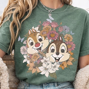 May include: Green t-shirt featuring Chip and Dale surrounded by a floral arrangement of pink, orange, and white flowers. The cartoon characters are smiling. Butterflies are also included in the design.