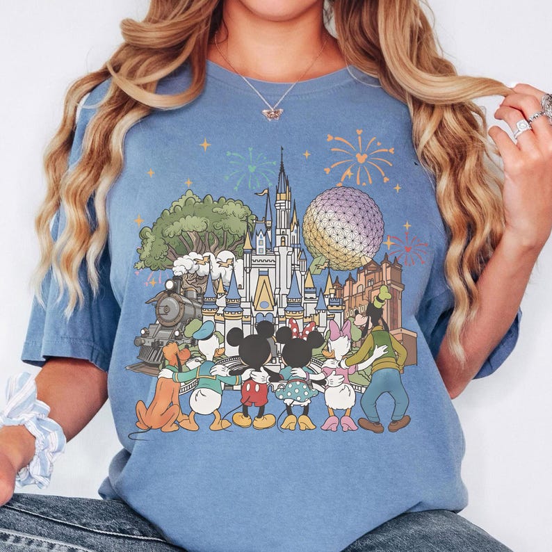 May include: A light blue t-shirt featuring a colorful graphic of a castle, a train, and various cartoon characters. The graphic includes fireworks and a large sphere. The characters are embracing each other. The shirt is a comfortable, casual style.