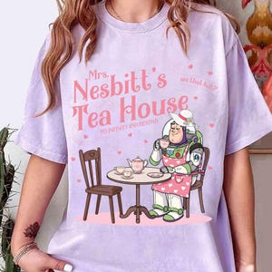 May include: A light purple t-shirt featuring a cartoon illustration of Buzz Lightyear in a tea party scene. The text "Mrs. Nesbitt's Tea House" is displayed in pink, with the phrase "To infinity and beyond" below.