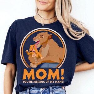 May include: Navy blue t-shirt featuring a cartoon lioness and cub within a circular design. The design includes the text "MOM! YOU'RE MESSING UP MY MANE!" in orange. The shirt is a comfortable, casual style.