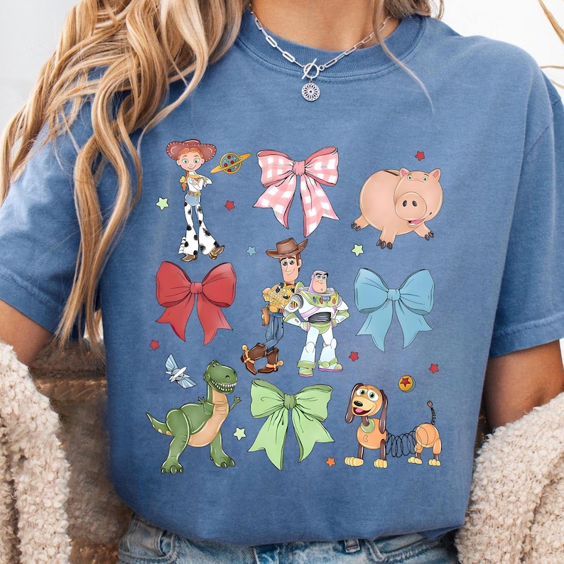 May include: Blue t-shirt featuring colorful cartoon illustrations of characters and bows. The design includes a pink pig, a green dinosaur, a dog with a spring body, and other characters. The shirt has a crew neck.