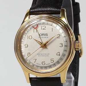 Może przedstawiać: Złoty zegarek na rękę Oris z białą tarczą, czarnymi cyframi i czarnym skórzanym paskiem. Zegarek posiada wyświetlacz daty oraz napisy "WATERPROOF ANTISHOCK". Wskazówki i indeksy są złote. Klasyczny styl.