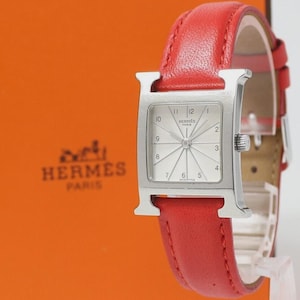 May include: A silver-toned watch with a rectangular face and a red leather band. The watch face has a radial design with silver hands and hour markers. The band is a vibrant red, and the watch is set against an orange background.