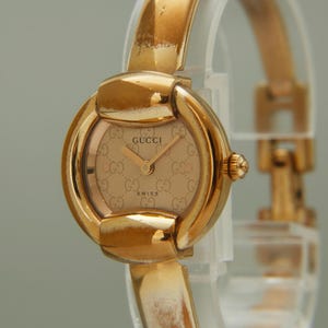 May include: A gold-tone Gucci wristwatch featuring a round case with a polished finish and a matching bangle-style bracelet. The dial displays a repeating GG logo pattern in a light gold hue, with the Gucci brand name and Swiss text printed.