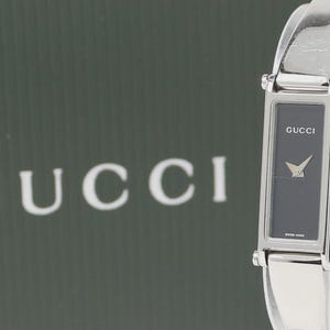 Może przedstawiać: Srebrny zegarek Gucci z prostokątną czarną tarczą. Zegarek ma srebrną bransoletę i złotą wskazówkę. Słowo "GUCCI" jest wydrukowane na tarczy zegarka. Tło przedstawia słowo "GUCCI" dużymi białymi literami.