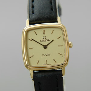 May include: A gold-toned Omega De Ville wristwatch with a square face and black leather band. The watch face is cream-colored with black hands and hour markers. The watch is marked "Omega" and "De Ville" with "Swiss Made" at the bottom.