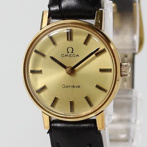 May include: A gold-toned Omega Genève wristwatch with a black leather band. The watch face is gold with black hands and markers. The Omega logo and "Genève" are printed on the face. The watch has a round face and a gold-colored case.