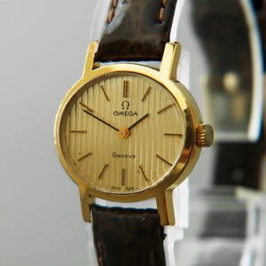 Pode incluir: Um relógio Omega Genève dourado com uma bracelete de couro castanho. O mostrador do relógio é dourado com linhas verticais e ponteiros pretos. O relógio tem o logótipo Omega e a palavra "Genève".
