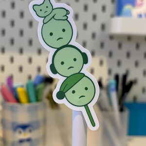Pode incluir: Um autocolante verde com tr&ecirc;s personagens de desenhos animados empilhadas. A personagem do topo &eacute; um gato, a do meio tem auscultadores e a de baixo usa um chap&eacute;u. Todos t&ecirc;m uma express&atilde;o triste. O autocolante est&aacute; numa caneta branca.