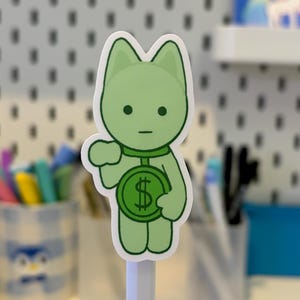 Pode incluir: Um autocolante verde com a forma de um gato a segurar uma moeda com um s&iacute;mbolo de d&oacute;lar. O autocolante tem uma borda branca e est&aacute; montado num l&aacute;pis branco. O fundo &eacute; uma parede perfurada branca.