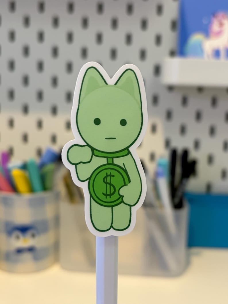 Pode incluir: Um autocolante verde com a forma de um gato a segurar uma moeda com um s&iacute;mbolo de d&oacute;lar. O autocolante tem uma borda branca e est&aacute; montado num l&aacute;pis branco. O fundo &eacute; uma parede perfurada branca.