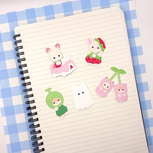 May include: A spiral-bound notebook with several colorful stickers. The stickers include a bunny in a pink car, a bunny in a strawberry dress, a green melon-themed character, a white ghost, and pink cherry-shaped characters.