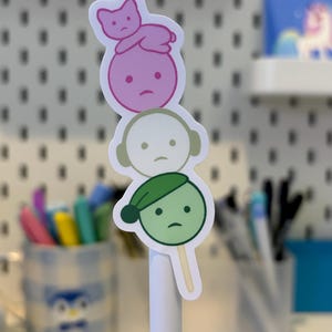 Pode incluir: Um adesivo com tr&ecirc;s personagens de desenho animado empilhados, cada um com uma express&atilde;o triste. O personagem superior &eacute; rosa com um gato, o do meio &eacute; branco com fones de ouvido e o inferior &eacute; verde com um chap&eacute;u.