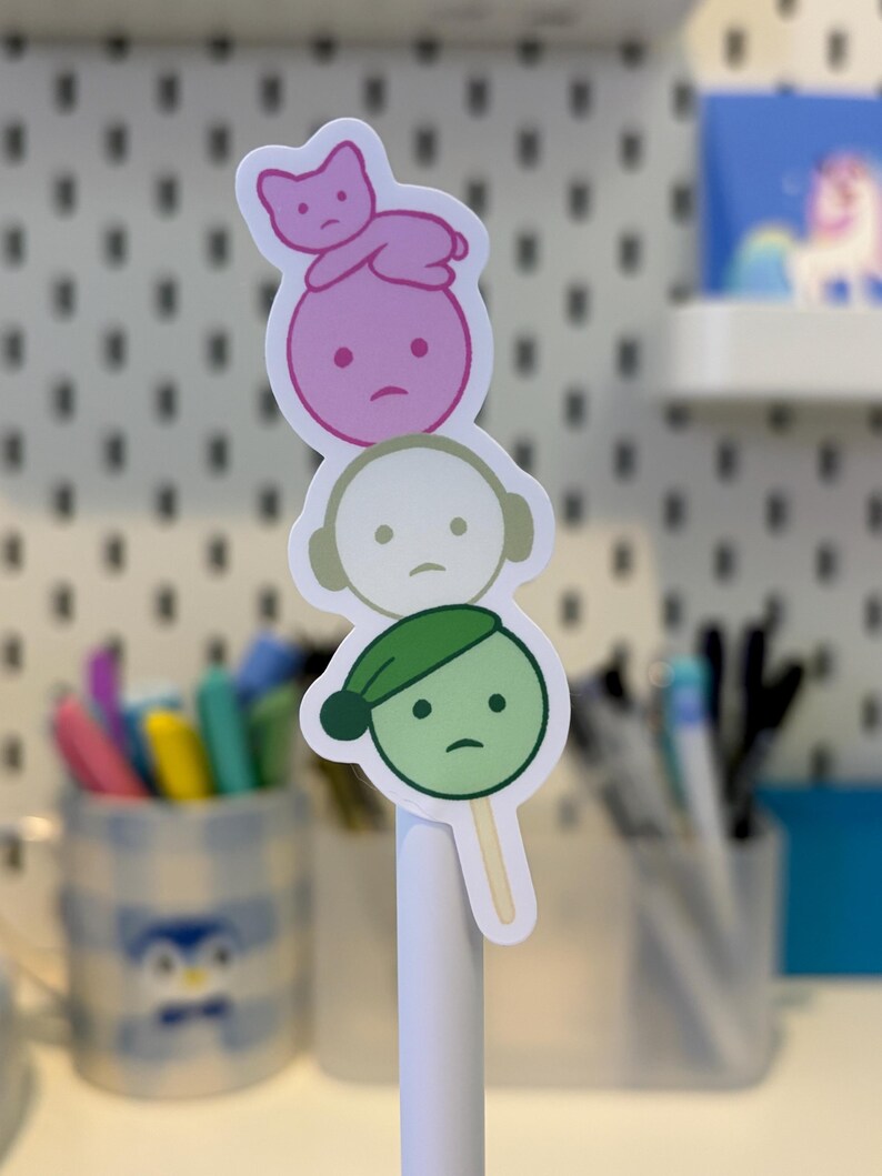 Pode incluir: Um adesivo com tr&ecirc;s personagens de desenho animado empilhados, cada um com uma express&atilde;o triste. O personagem superior &eacute; rosa com um gato, o do meio &eacute; branco com fones de ouvido e o inferior &eacute; verde com um chap&eacute;u.