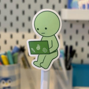 Pode incluir: Um autocolante verde de uma personagem de desenho animado a usar um computador port&aacute;til. A figura tem uma cabe&ccedil;a e um corpo redondos, com uma cara simples. O ecr&atilde; do port&aacute;til tem pequenos autocolantes. O autocolante est&aacute; colado a um l&aacute;pis branco.