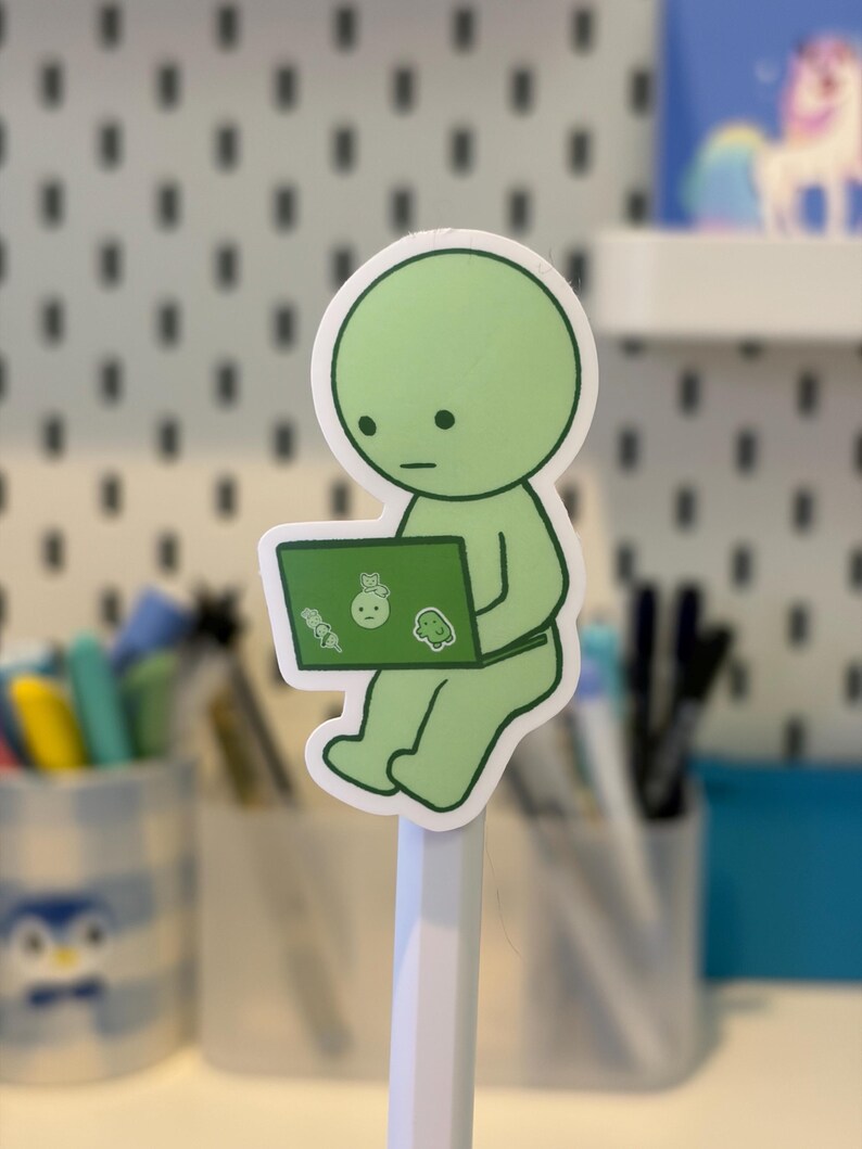 Pode incluir: Um autocolante verde de uma personagem de desenho animado a usar um computador port&aacute;til. A figura tem uma cabe&ccedil;a e um corpo redondos, com uma cara simples. O ecr&atilde; do port&aacute;til tem pequenos autocolantes. O autocolante est&aacute; colado a um l&aacute;pis branco.