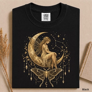 May include: A black t-shirt featuring a gold graphic of a fairy with wings sitting on a crescent moon, surrounded by stars and hanging ornaments. Below the moon is a large moth with intricate wing patterns. The neck label reads Comfort Colors.