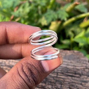 May include: Two silver spiral rings, each with multiple bands. The rings are shown against a blurred backdrop of green foliage and a dark wooden surface. The rings are designed to wrap around the finger.