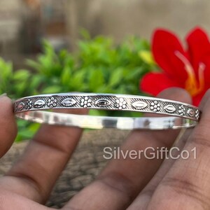 May include: A silver bangle bracelet with an intricate pattern of oval and floral designs. The bracelet is displayed against a backdrop of green foliage and a red flower. The bracelet is approximately 0.25 inches wide.