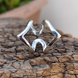 Könnte beinhalten: Ein silberfarbener Ring mit einem offenen, geometrischen Design, scharfen, kantigen Ecken und glatten, geschwungenen Abschnitten. Die Metalloberfläche ist poliert und reflektierend, was eine markante, facettierte Form erzeugt.