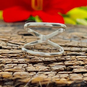 Può includere: Un anello color argento con un design a forma di X. L'anello è posto su una superficie di legno marrone scuro strutturata, con un fiore rosso sfocato e fogliame verde sullo sfondo. Il design dell'anello presenta due fasce che si incrociano.