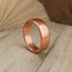 May include: A copper-colored ring with a smooth, polished finish. The ring has a rounded band and a simple, elegant design. The ring is displayed on a light-colored wooden surface.