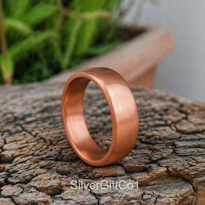 Può includere: Un anello in rame massiccio con una finitura spazzolata liscia e un profilo arrotondato. La fascia presenta un caldo colore bruno-rossastro metallico e una larghezza costante. Il testo SilverGiftCo1 è visibile nella parte inferiore.