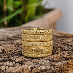 Könnte beinhalten: Ein Stapel von sechs goldfarbenen Ringen mit aufwendigen floralen und geometrischen Mustern. Die Ringe sind auf einem Stück strukturiertem Holz gestapelt, mit einem verschwommenen grünen Hintergrund. Die Ringe haben eine Vintage-Ästhetik.