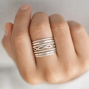 Peut inclure: Un empilement de six bagues en argent portées sur un doigt. L'ensemble comprend deux anneaux lisses, deux anneaux à texture torsadée, un anneau avec un motif de maillons infinis délicats et un anneau lisse supplémentaire, tous avec une finition polie.