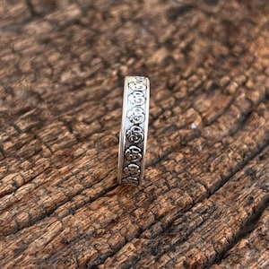 May include: A silver ring with a repeating floral pattern. The ring is set against a rustic wooden surface, highlighting its intricate design. The ring's details are clearly visible, showcasing the craftsmanship.