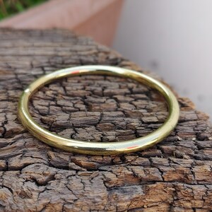 May include: A gold-colored, circular bangle bracelet. The smooth, polished metal reflects light, creating highlights. The bracelet is resting on a piece of weathered, dark brown wood with a textured surface. The bangle is a simple, elegant piece of jewelry.