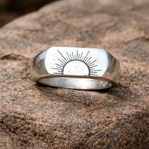 Puede incluir: Un anillo plateado con una cara rectangular con un diseño de sol. El anillo está sobre una superficie de piedra marrón texturizada. El diseño del sol consiste en un semicírculo con líneas radiantes.