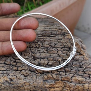 Könnte beinhalten: Ein silbernes Armreif-Armband mit einem schlichten, eleganten Design. Das Armband ist eine durchgehende Schlaufe aus poliertem Metall mit einem gewickelten Detail in der Nähe des Verschlusses. Der Armreif liegt auf einem Stück verwittertem Holz.