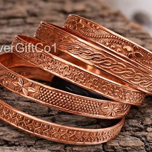 May include: Several copper bangles with detailed floral and geometric patterns are displayed on a textured wooden surface. The bangles have a polished finish and different designs. The text "SilverGiftCo1" is visible.