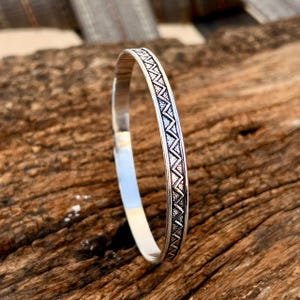 Op de afbeelding: Een zilveren armband met een geometrisch patroon. De armband heeft een gepolijste afwerking en een herhalend ontwerp van driehoeken en zigzags rond de band. De armband wordt gepresenteerd op een houten oppervlak.