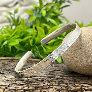 Könnte beinhalten: Ein silbernes Armreif-Armband mit gehämmertem Muster. Das offene Armband liegt auf einer Holzoberfläche neben einem Stein und grünem Laub. Das Armband trägt den Text "SilverGiftCo1".