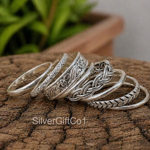Op de afbeelding: Een verzameling zilveren ringen met verschillende ontwerpen, waaronder bloemen-, gevlochten en patroonbanden. De ringen worden gepresenteerd op een houten oppervlak, met de tekst "SilverGiftCo1" zichtbaar.