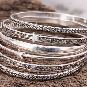 May include: A stack of silver bangles with various designs. Some bangles have a braided pattern, others feature a floral design with star accents. The bangles are displayed on a textured, brown surface.