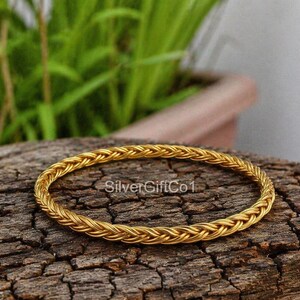 Può includere: Un braccialetto intrecciato color oro. Il braccialetto è circolare e realizzato con fili fittamente intrecciati, creando un aspetto strutturato. Il braccialetto è esposto su una superficie di legno scuro e strutturato, con fogliame verde sullo sfondo.