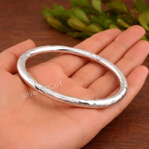 May include: A silver-coloured, hammered-finish bangle bracelet. The oval-shaped bracelet is held in a hand, with the text "SilverGiftCo1" visible. The bracelet has a textured surface and a simple, elegant design.