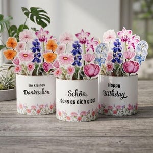 May include: Three cylindrical white containers featuring colorful watercolor-style floral arrangements on top. The containers display the German text "Ein kleines Dankeschön," "Schön, dass es dich gibt!," and the English text "Happy Birthday" with floral borders.