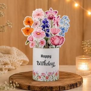Könnte beinhalten: Eine Pop-up-Grußkarte mit einem lebendigen Aquarell-Blumenstrauß in Orange, Pink, Violett und Blau, der aus einer weißen zylindrischen Basis hervorgeht. Die Basis zeigt den schwarzen Text Happy Birthday und eine zarte Blumenbordüre am unteren Rand.